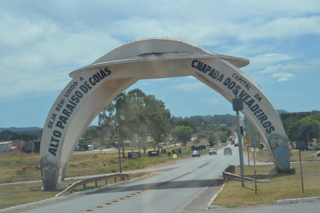 Concrete entrance arch above a paved road into Alto Paraíso de Goiás, with cars, trees and buildings beyond