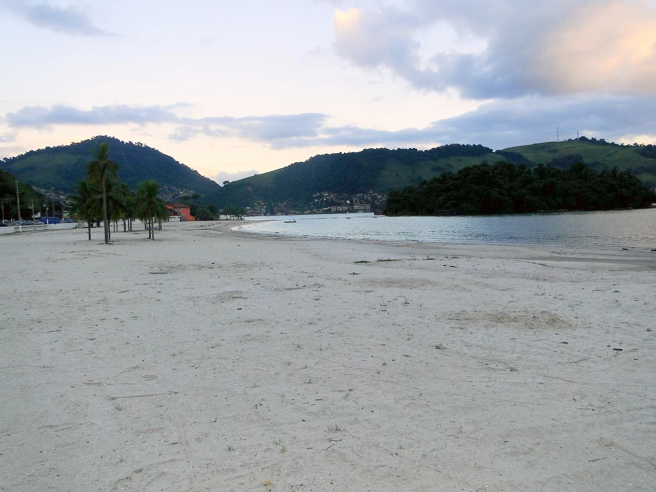 Broad sandy beach with palm trees beside calm bay water, wooded hills and shoreline buildings at Praia do Anil