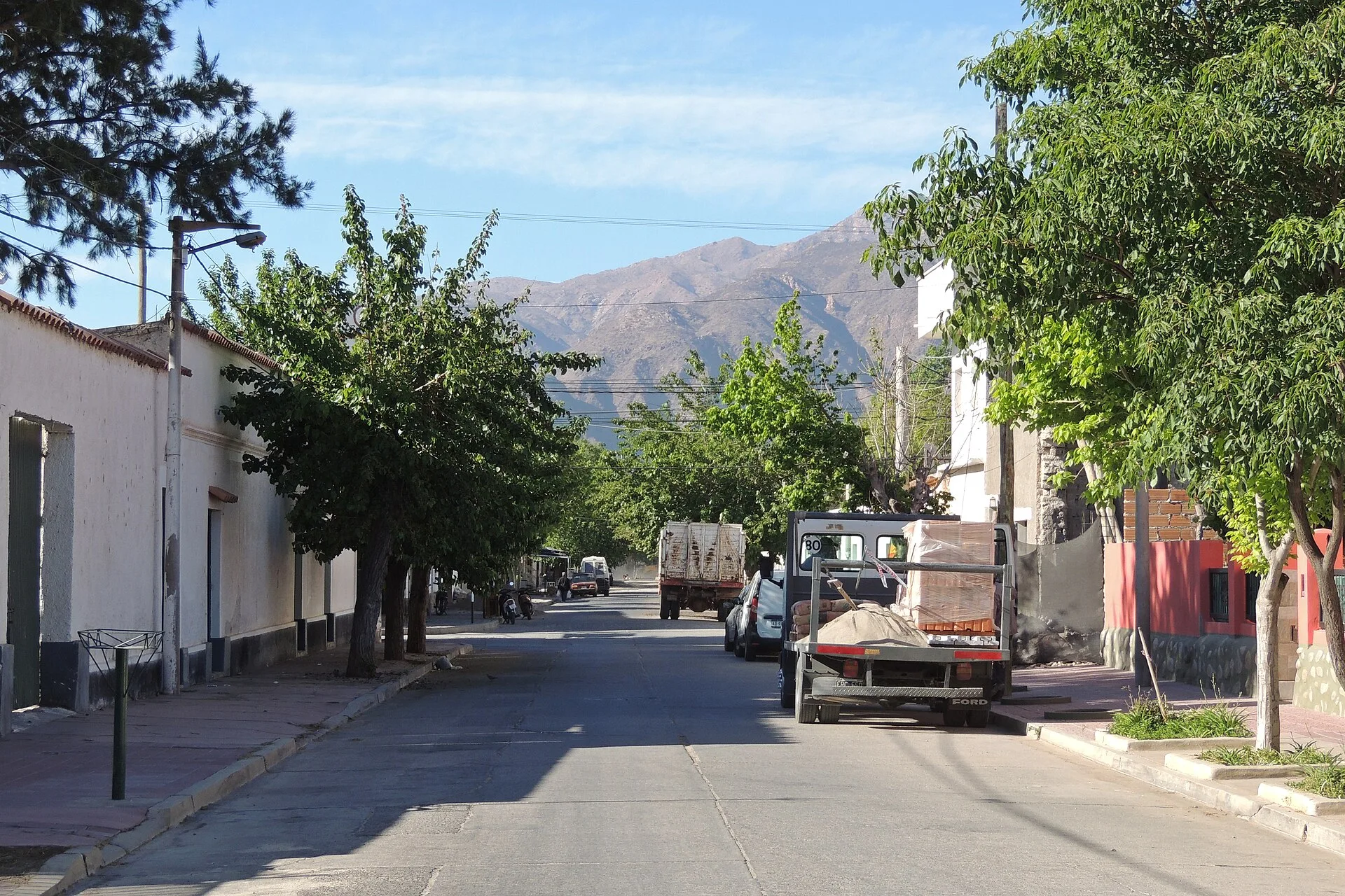 A Cafayate street with low buildings, trees, parked vehicles and mountains beyond