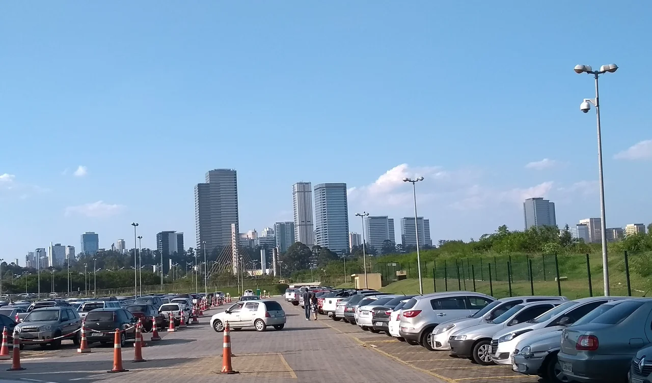 Parking and a footbridge in front of Alphaville high-rises under a partly cloudy sky