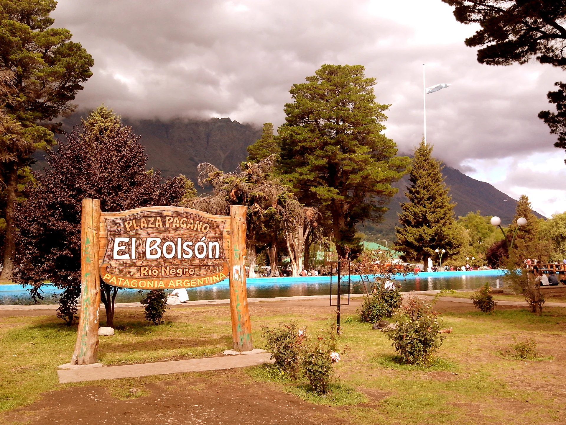 The wooden Plaza Pagano sign beside trees and the pond in El Bolsón