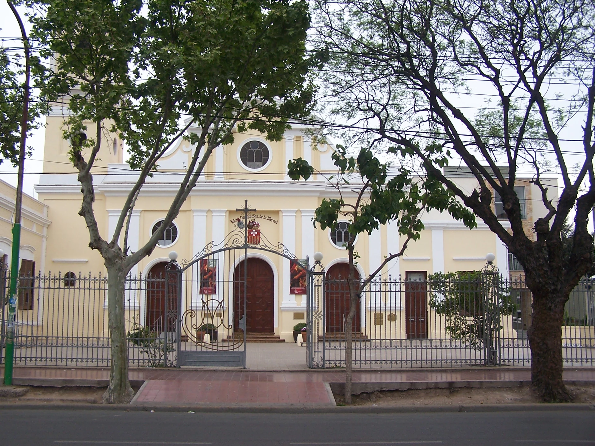 Pale yellow La Merced church behind trees and an iron fence in Maipú