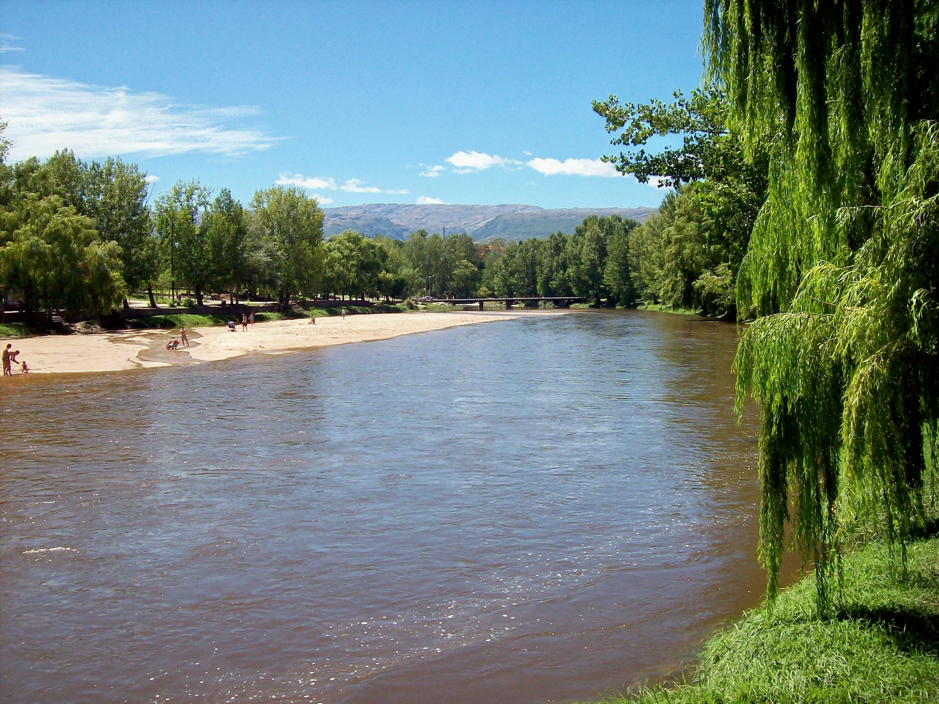 The Mina Clavero river beside trees and a sandy bank, with hills and a bridge in the distance