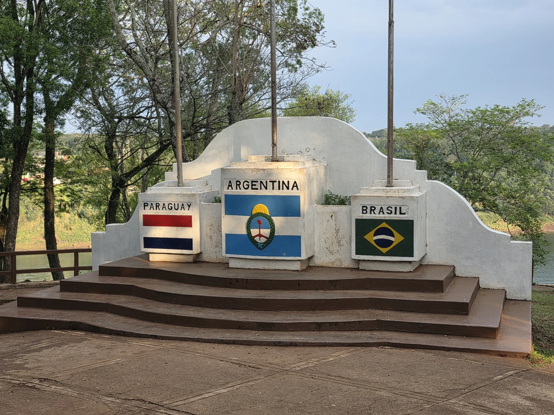 A monument at Hito Tres Fronteras in Puerto Iguazú with Argentina, Paraguay and Brazil emblems, trees and water behind it