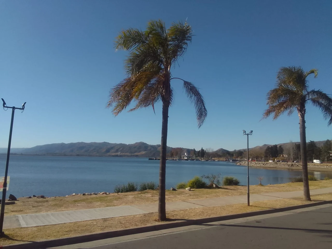 Palm trees beside a paved waterfront path, San Roque Lake and distant hills in Villa Carlos Paz