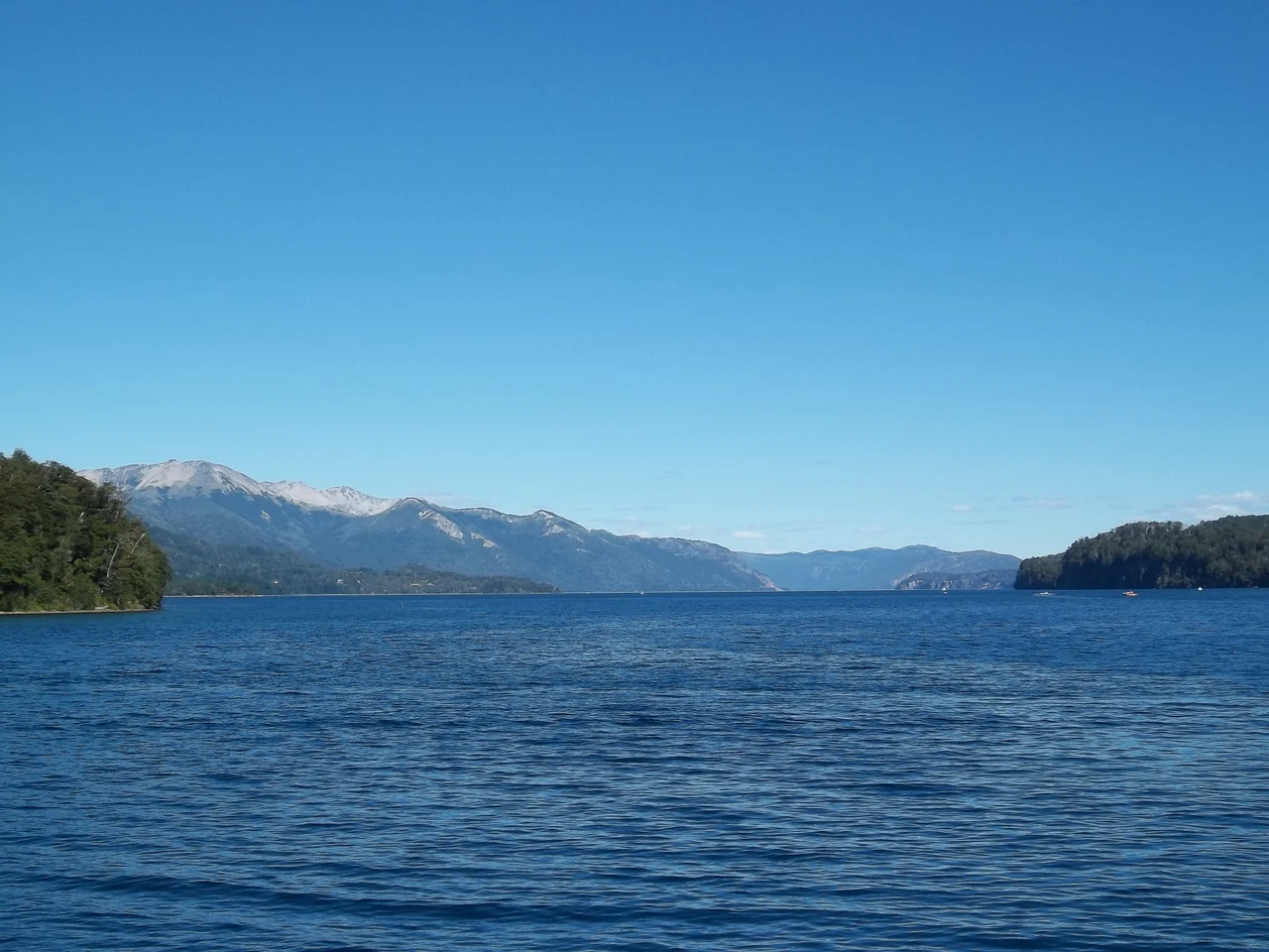 Blue Nahuel Huapi lake with forested shores, mountains and clear sky, viewed from the Villa La Angostura pier